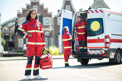 Smiling contented young female paramedic holding a medical bag and waiting for her colleagues outdoors