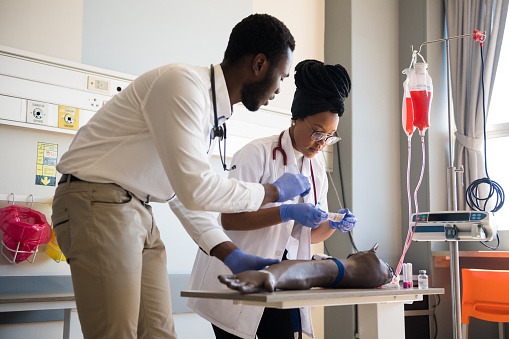 ClinicalPractice4 Young male and female doctors practicing IV Drip on artificial limb on desk. Healthcare colleagues are in training class. They are in hospital.